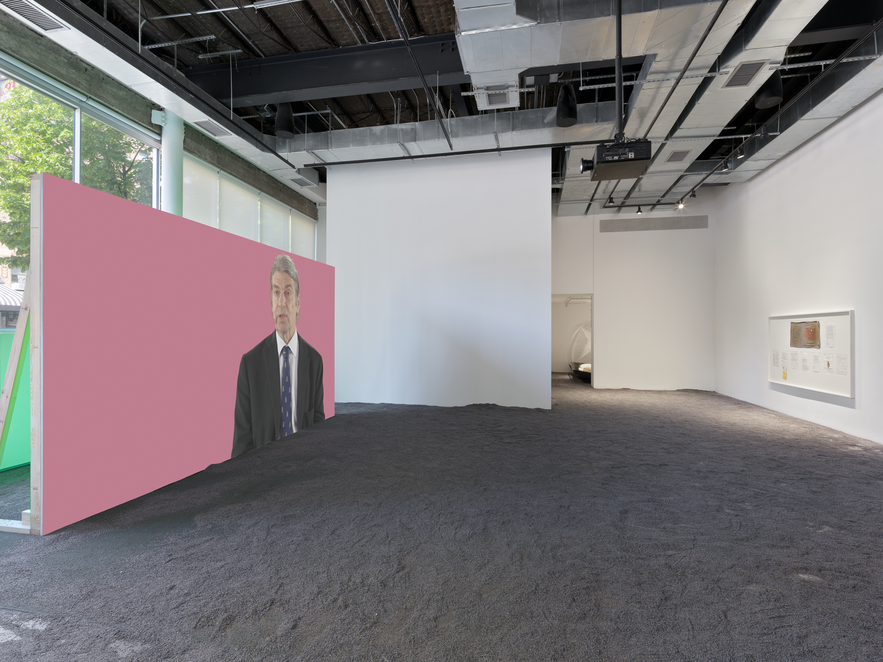 Projected image of a man in a suit against a pink background beside a white-framed display of exhibition documents; lavender flooring throughout.