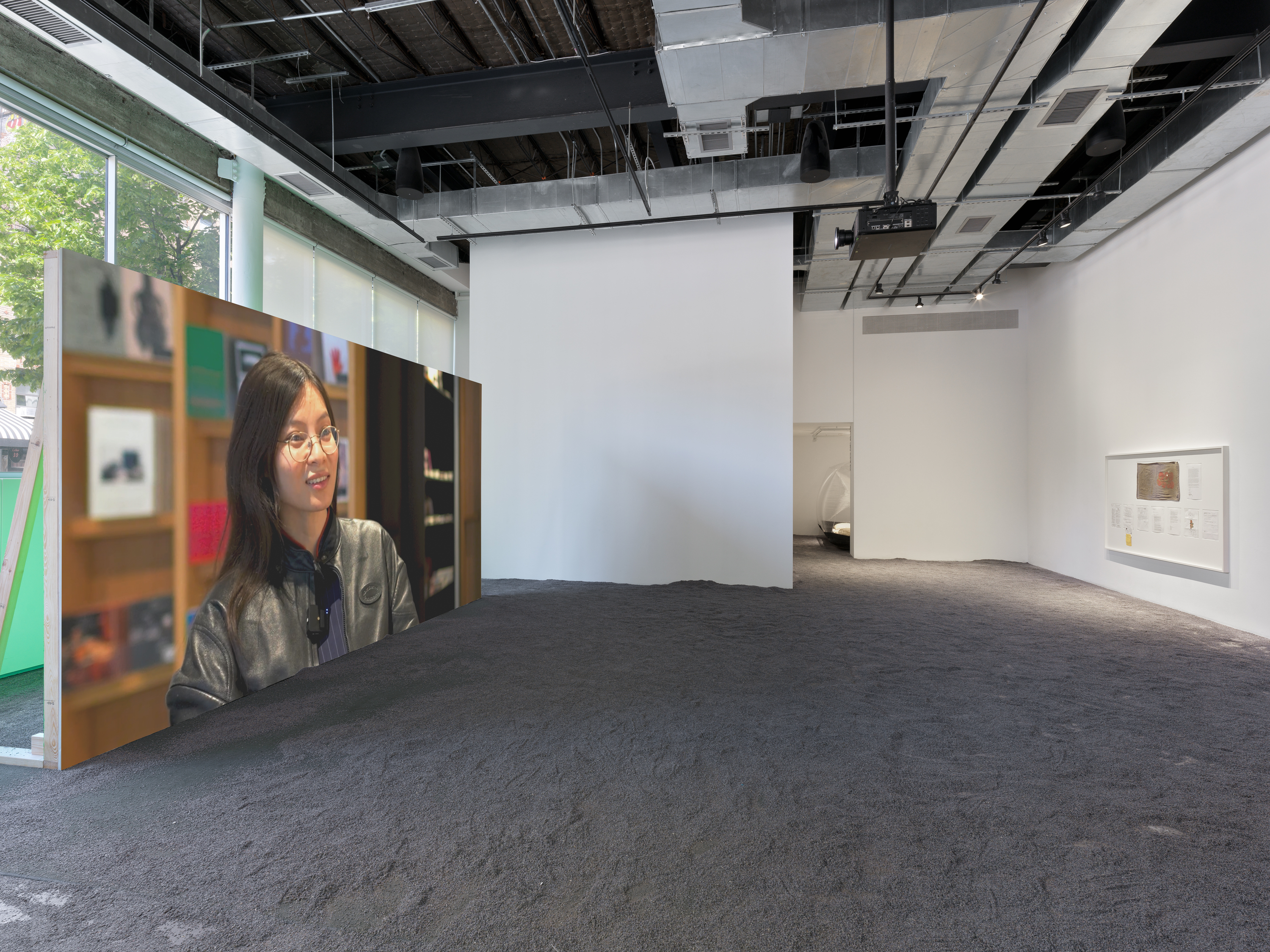 Projected image of a woman wearing glasses seated before a bookshelf beside a white-framed display of exhibition documents; lavender flooring throughout.