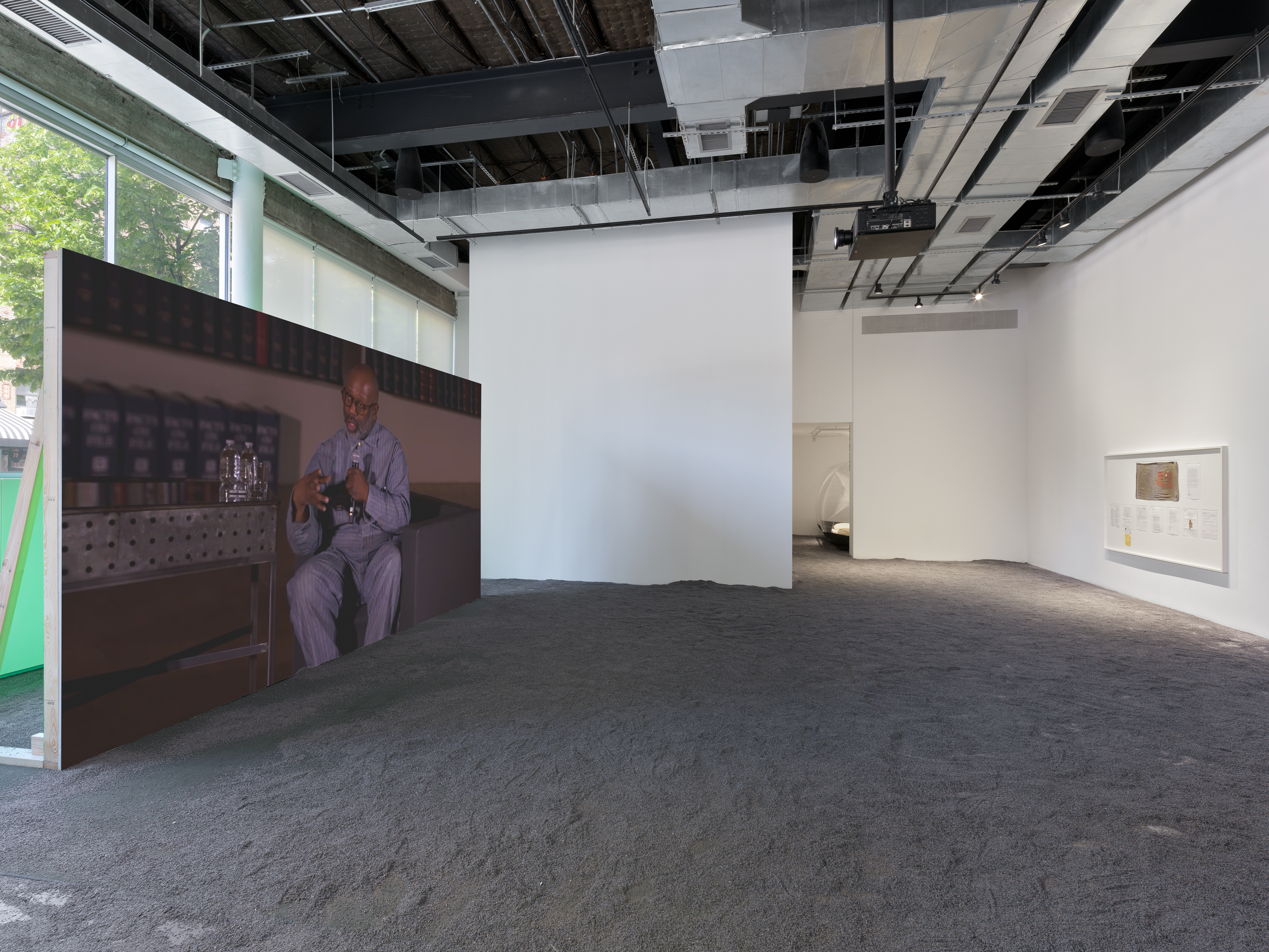 Projected image of a man in a gray pinstriped suit speaking into a microphone with two water bottles in front of a bookshelf; white-framed exhibition documents displayed nearby on lavender flooring.