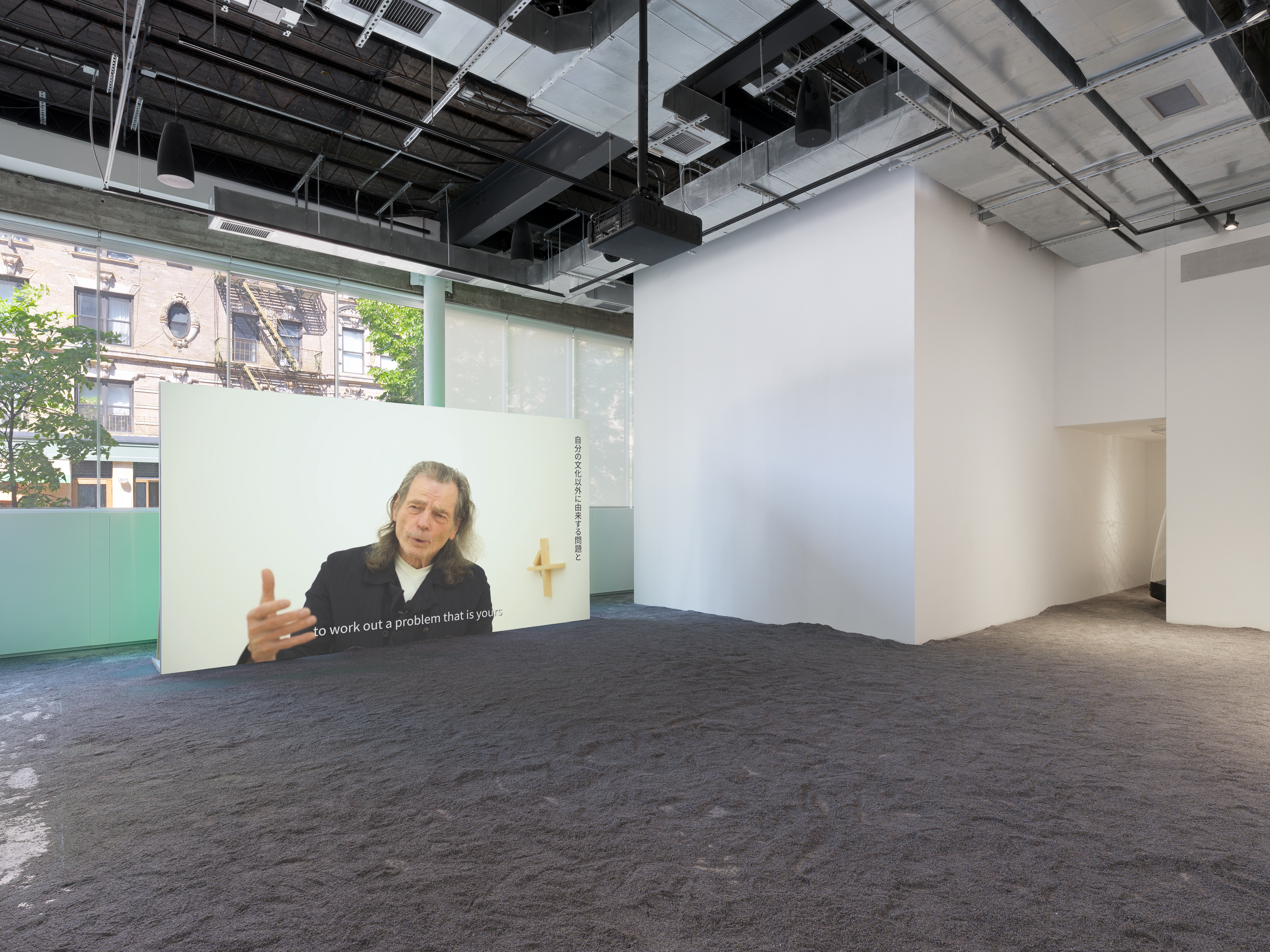 Projected image of a man in a black shirt layered over a white undershirt speaking beside Japanese kanji text; lavender flooring throughout.