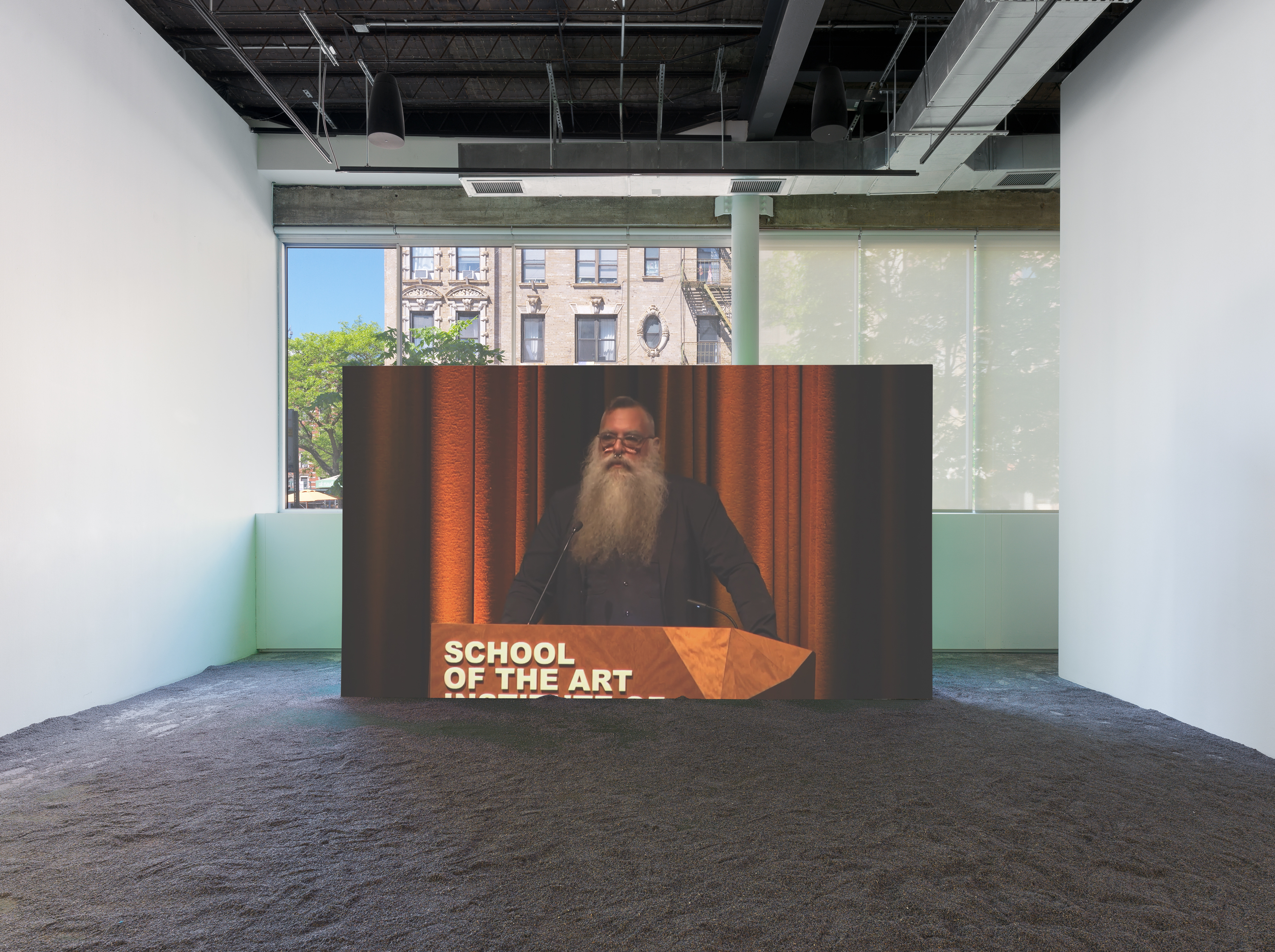 A bearded man wearing glasses speaks at a podium with the words “SCHOOL OF THE ART” projected onto a wooden structure; lavender flooring throughout.