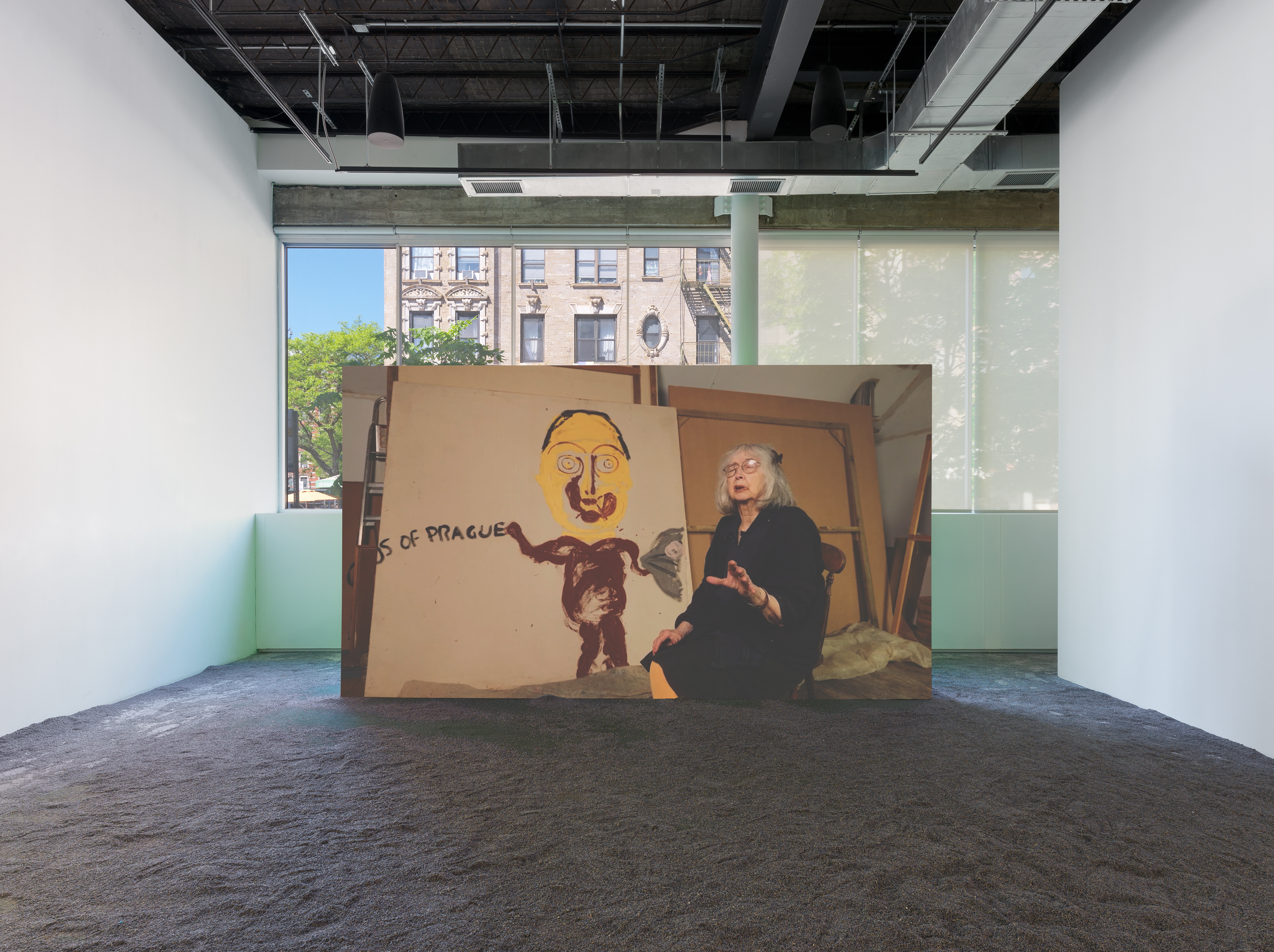A woman wearing glasses sits before a projected artwork depicting a humanoid figure with a yellow face and brown body beside the word “PRAGUE”; lavender flooring throughout.