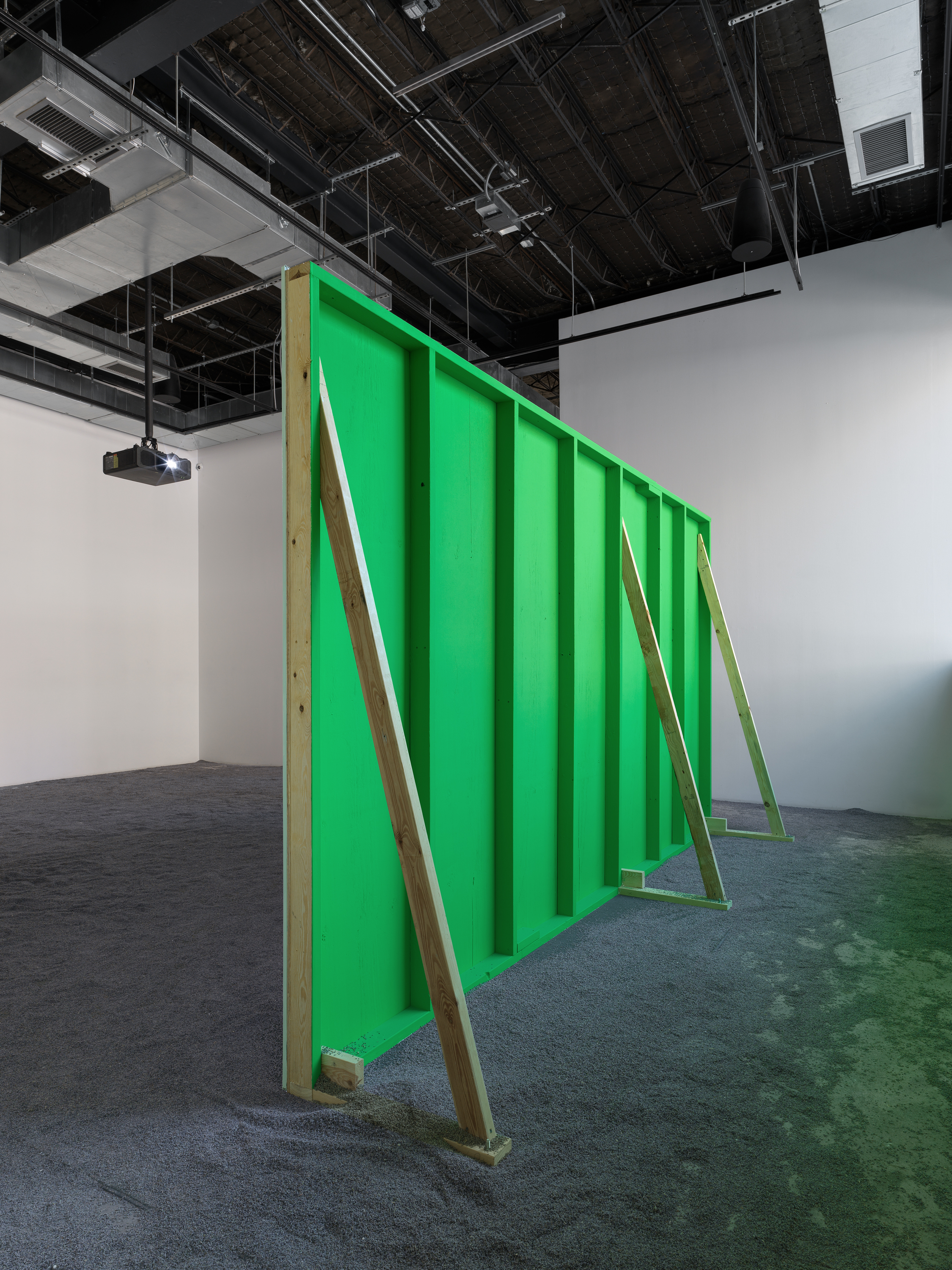 Rear view of a green-painted wooden projection structure supported by three poles with a projector nearby; lavender flooring throughout.