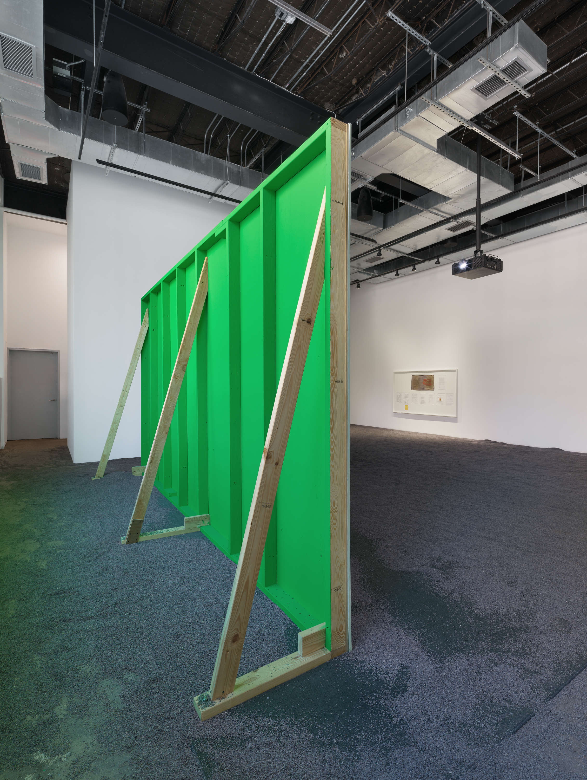 Rear view of a green-painted wooden projection structure supported by three poles with a projector nearby and a white-framed display of exhibition documents on the right; lavender flooring throughout.